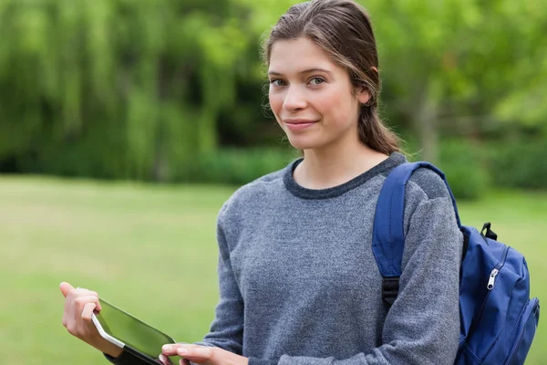 Smiling young woman holding a tablet pc while standing in a park — Stock Photo, Image