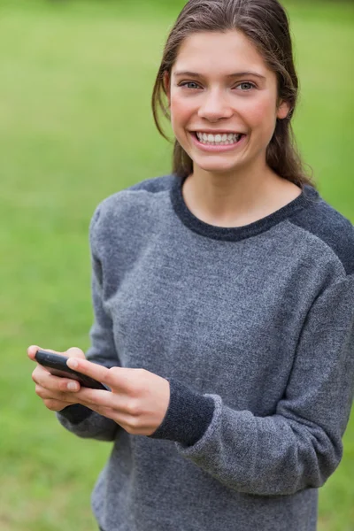 Junges lächelndes Mädchen, das in die Kamera schaut, während sie ihr Handy benutzt — Stockfoto
