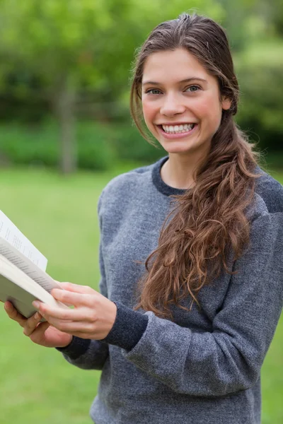 Joven chica sonriente mirando a la cámara mientras lee un libro —  Fotos de Stock
