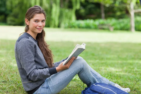 Chica sonriente sosteniendo un libro de rodillas mientras está sentada en el gr —  Fotos de Stock