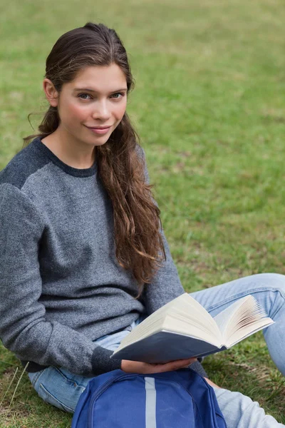 Estudiante tranquila sosteniendo un libro mientras está sentada junto a su mochila —  Fotos de Stock