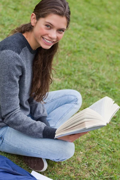 Young smiling woman sitting cross-legged in a park while holding — Stock Photo, Image