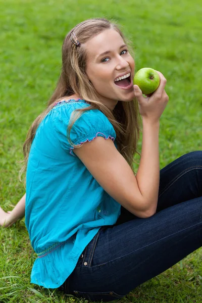 Молода усміхнена жінка їсть зелене яблуко, сидячи в парку — стокове фото