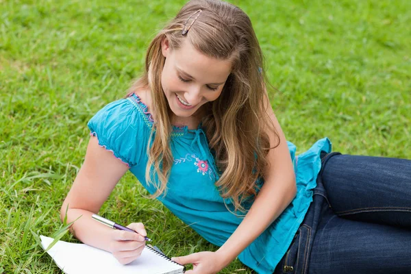 Junges lächelndes Mädchen, das auf dem Gras liegt, während es auf ein Notizbuch schreibt — Stockfoto