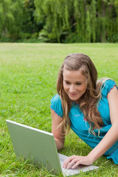 Adolescente sonriente acostada en un parque mientras escribe en su computadora portátil —  Fotos de Stock