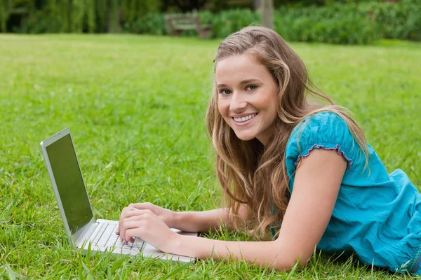 Smiling teenager using her laptop while lying down in a park — Stock Photo, Image