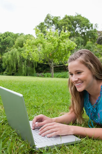 Joven chica sonriente usando su computadora portátil en un parque mientras está acostada en el —  Fotos de Stock