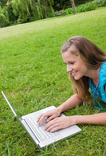 Lächelnde junge Frau, die im Park liegt, während sie an ihrem Laptop arbeitet — Stockfoto