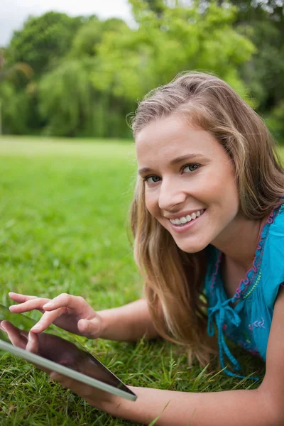 Mujer joven sonriente acostada en un parque mientras usa su ficha — Foto de Stock