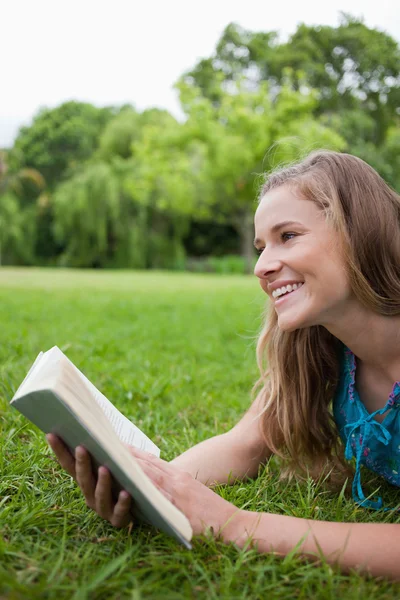Unga leende kvinna ligger i ett parklandskap medan du håller en bok — Stockfoto