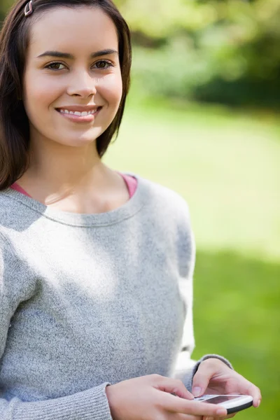 Sorrindo jovem mulher usando seu celular enquanto estava de pé em um parque — Fotografia de Stock