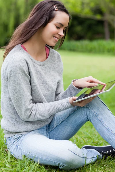 Young smiling woman sitting on the grass in a park while using h — Stock Photo, Image