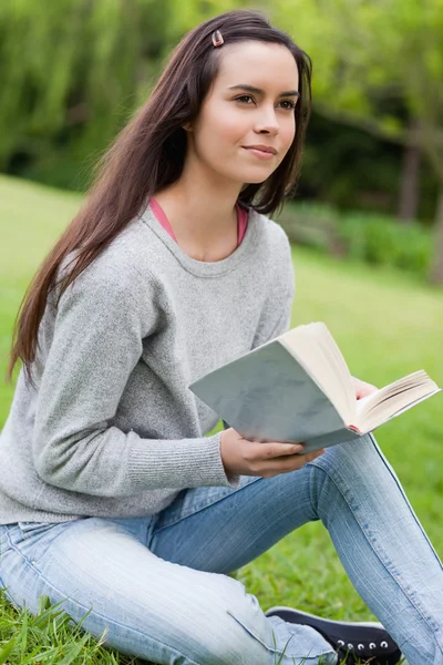 Mujer joven y pensativa sosteniendo un libro mientras está sentada en la hierba —  Fotos de Stock