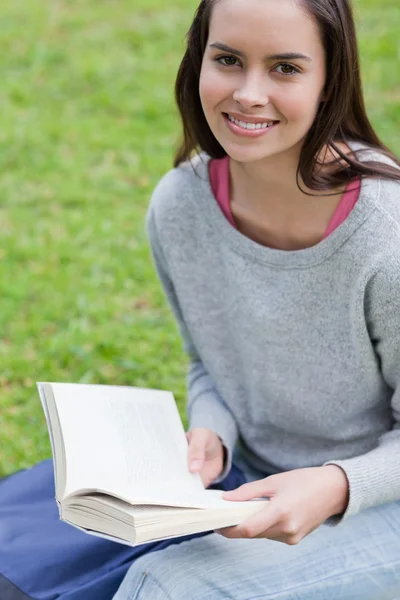 Jovem mulher sorrindo lendo um livro em um parque enquanto olha para o th — Fotografia de Stock