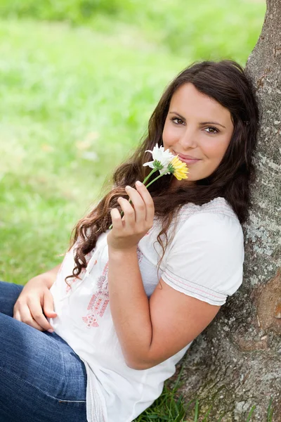 Unga leende kvinna att lukta blommor sittande mot en tre — Stockfoto