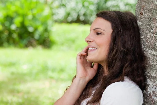 Young smiling woman leaning against a tree while talking on the — Stock Photo, Image