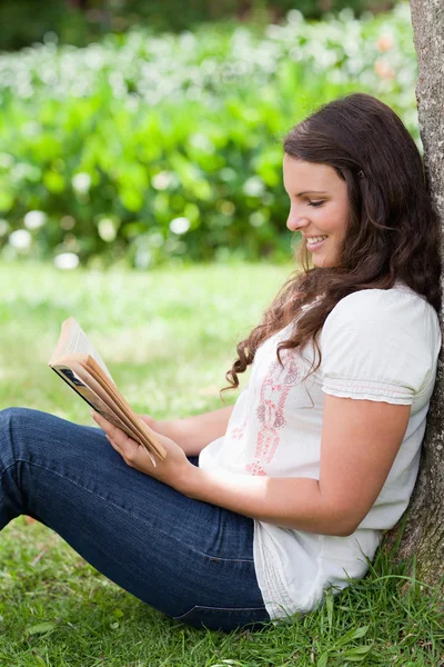 Young smiling woman reading a book while leaning against a tree — Stock Photo, Image
