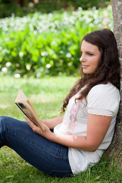Joven chica reflexiva apoyada contra un árbol mientras lee un boo —  Fotos de Stock