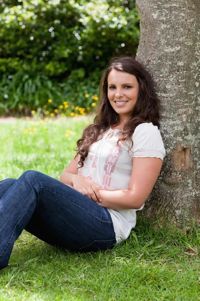Mladá usměvavá žena sedící proti strom s rukama přes — Stock fotografie