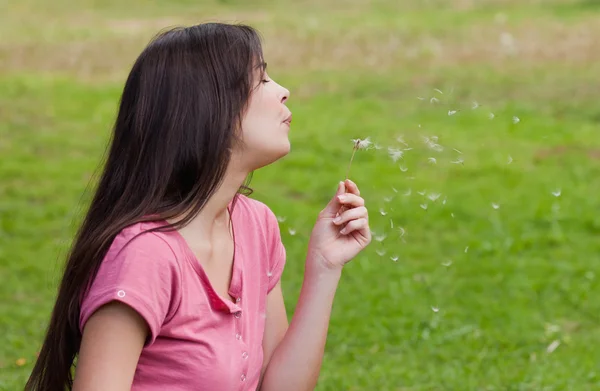 Young woman standing up in a park while blowing a dandelion — Stock Photo, Image
