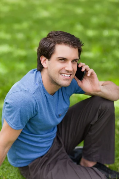 Młody człowiek szczęśliwy, za pomocą swojego telefonu komórkowego siedząc na gras — Zdjęcie stockowe