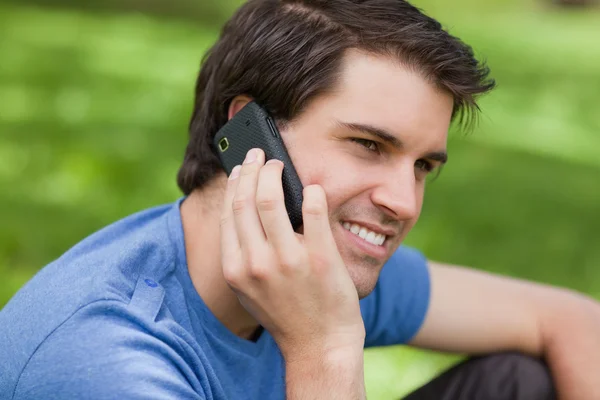 Smiling young man sitting in the countryside while using his cel — Stockfoto