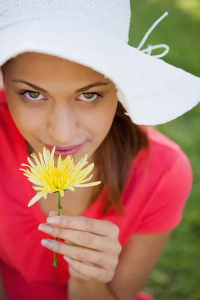 Mulher usando um chapéu branco enquanto cheira uma flor enquanto olha — Fotografia de Stock