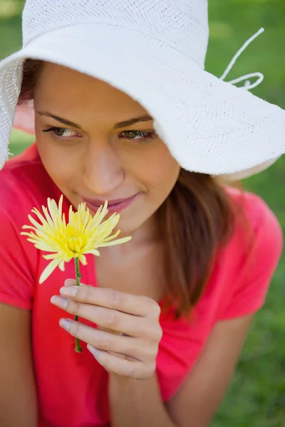 Mulher usando um chapéu branco enquanto cheira uma flor enquanto olha — Fotografia de Stock