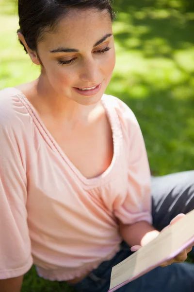 Γυναίκα που είναι χαμογελώντας διαβάζοντας ένα βιβλίο, όπως η ίδια κάθεται στο γρασίδι — Φωτογραφία Αρχείου