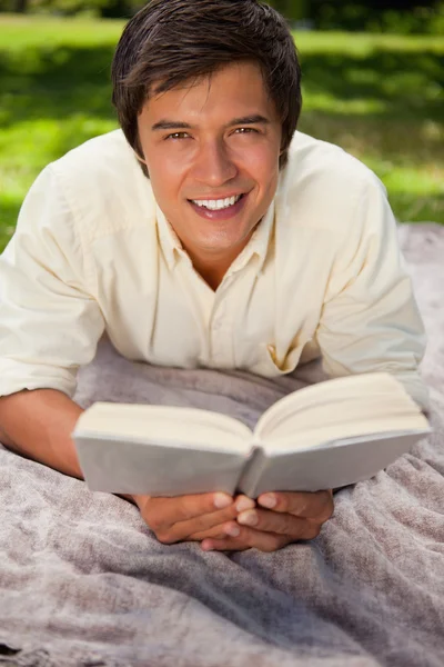 Człowiek uśmiechający się czytając książkę, jak on leży na kocu — Zdjęcie stockowe