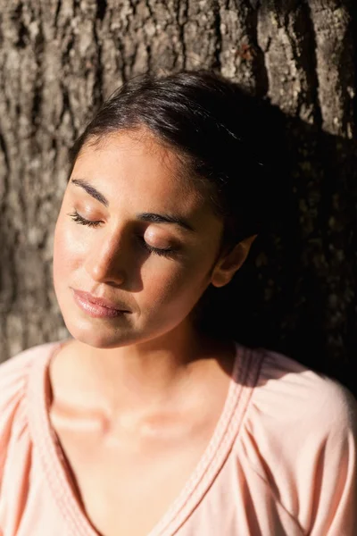 Mulher fechando os olhos enquanto sentada contra uma árvore — Fotografia de Stock