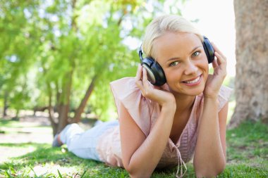 Smiling woman listening to music while lying on the lawn clipart