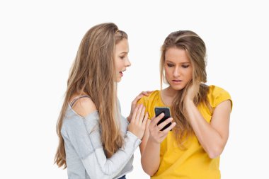 Upset young woman holding her cellphone consolded by her friend clipart