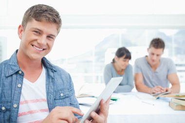 A smiling student using his tablet with his friends in the backg clipart