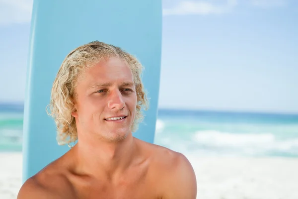 Jovem sorridente sentado na praia com sua prancha de surf — Fotografia de Stock