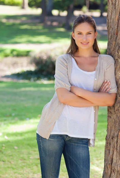 Mulher sorridente com os braços cruzados encostada a uma árvore — Fotografia de Stock