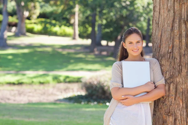 Femme souriante avec un ordinateur portable appuyé contre un arbre — Photo