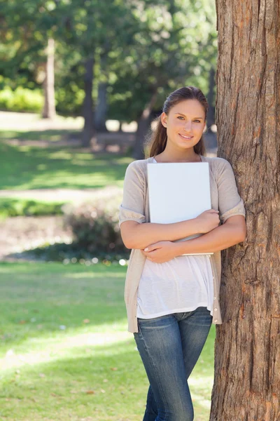 Smiling woman holding a laptop while leaning against a tree — Stock Photo, Image