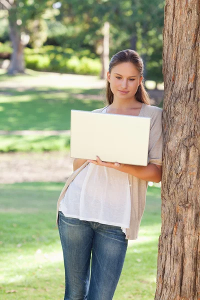 Mulher usando um laptop enquanto se inclina contra uma árvore — Fotografia de Stock