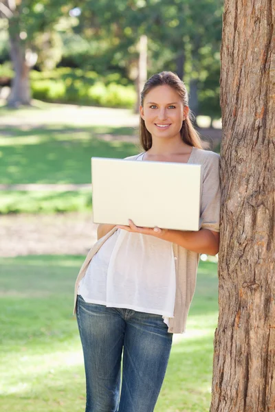 Smiling woman using a laptop while leaning against a tree — Stock Photo, Image