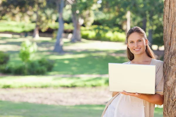 Smiling woman using a notebook while leaning against a tree — Stock Photo, Image