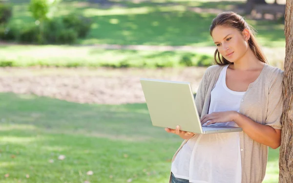 Woman leaning against a tree while using a laptop — Stock Photo, Image