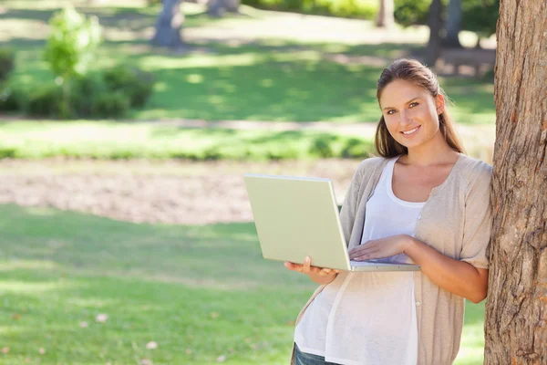 Smiling woman with a notebook leaning against a tree — Stock Photo, Image