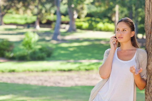 Frau telefoniert mit ihrem Handy, während sie sich gegen einen Baum lehnt — Stockfoto