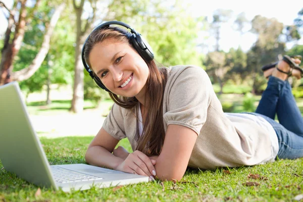 Mulher sorrindo com um fone de ouvido e um laptop deitado no gramado — Fotografia de Stock