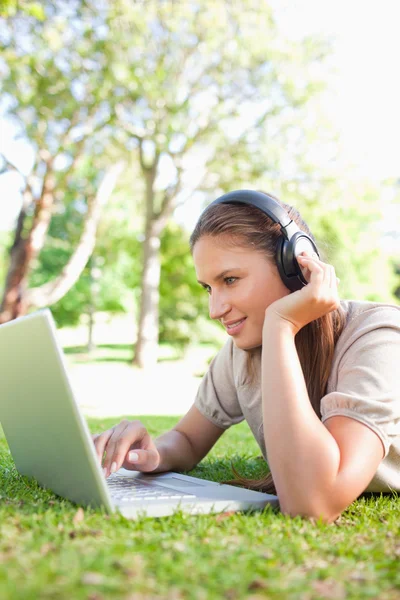 Mulher com fones de ouvido e um laptop deitado no gramado — Fotografia de Stock