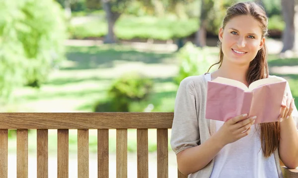 Mulher sorridente lendo um livro em um banco de parque — Fotografia de Stock