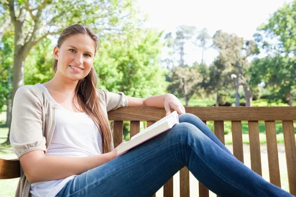 Mulher sorridente sentada em um banco com um livro — Fotografia de Stock