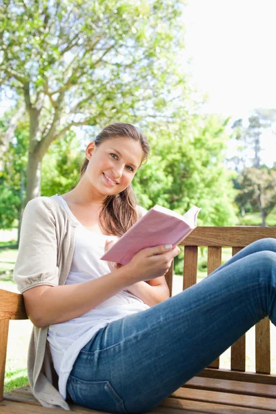 Χαμογελαστή γυναίκα που κάθεται σε ένα παγκάκι στο πάρκο με ένα βιβλίο — Φωτογραφία Αρχείου