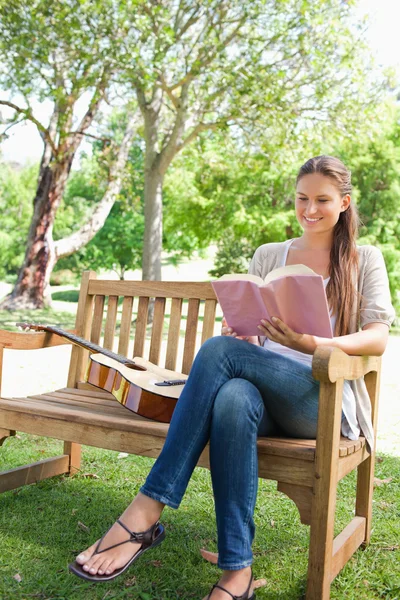 Mulher sorridente sentada em um banco com um livro e uma guitarra — Fotografia de Stock